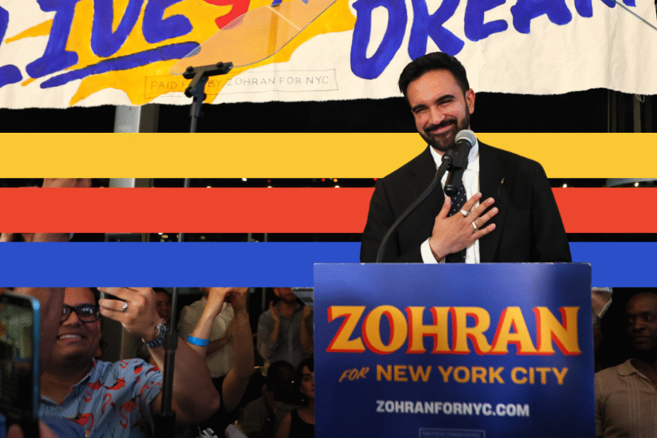 Color image of a mixed-race man with dark hair and a goatee in a suit with his hand over his heart at a podium featuring a banner reading "Zohran for New York City"