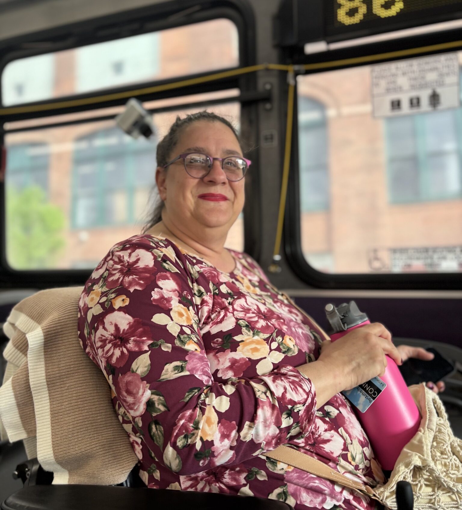 A person in a red floral dress and power chair smiles aboard a city bus