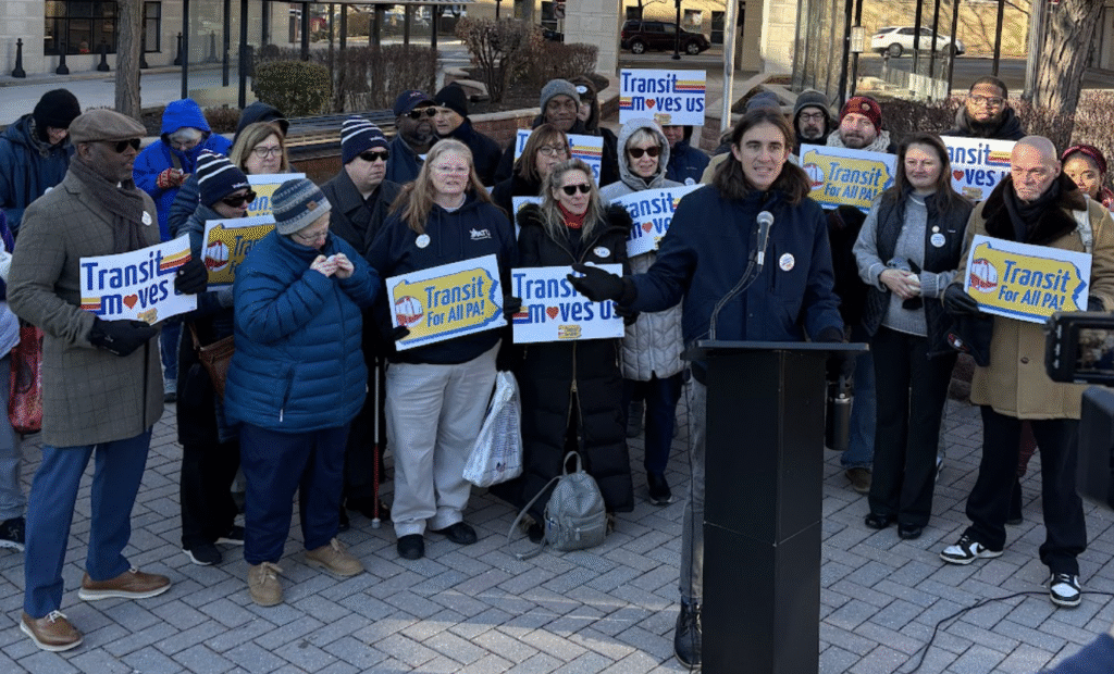 Color image of a multiracial, multi-age group of transit advocates holding signs that read "Transit Moves Us" and "Transit For All PA" behind a person at a podium speaking with their hands out